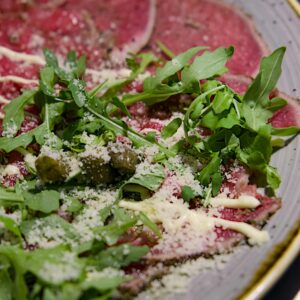 A close-up of fresh beef carpaccio garnished with arugula leaves and grated Parmesan cheese.